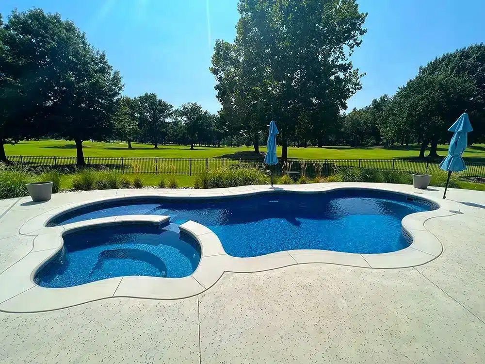 Freeform backyard swimming pool with attached spa, blue water, concrete deck, and green landscape view
