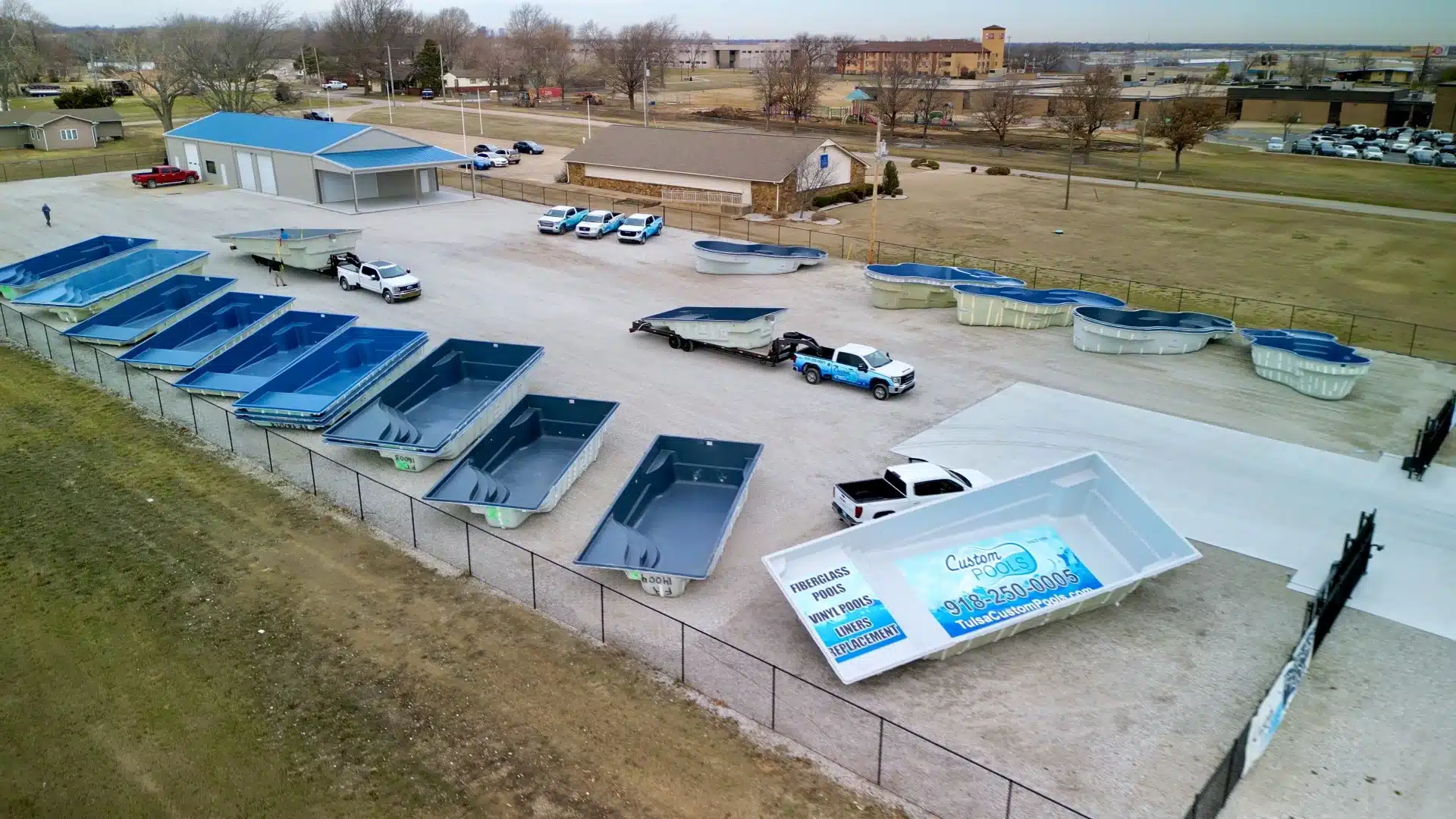 Aerial view of fiberglass swimming pools lined up in a storage yard with trucks and installation facility