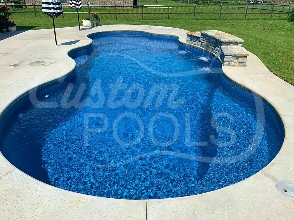 Freeform swimming pool with clear blue water, built-in waterfall feature, concrete deck, and stone design