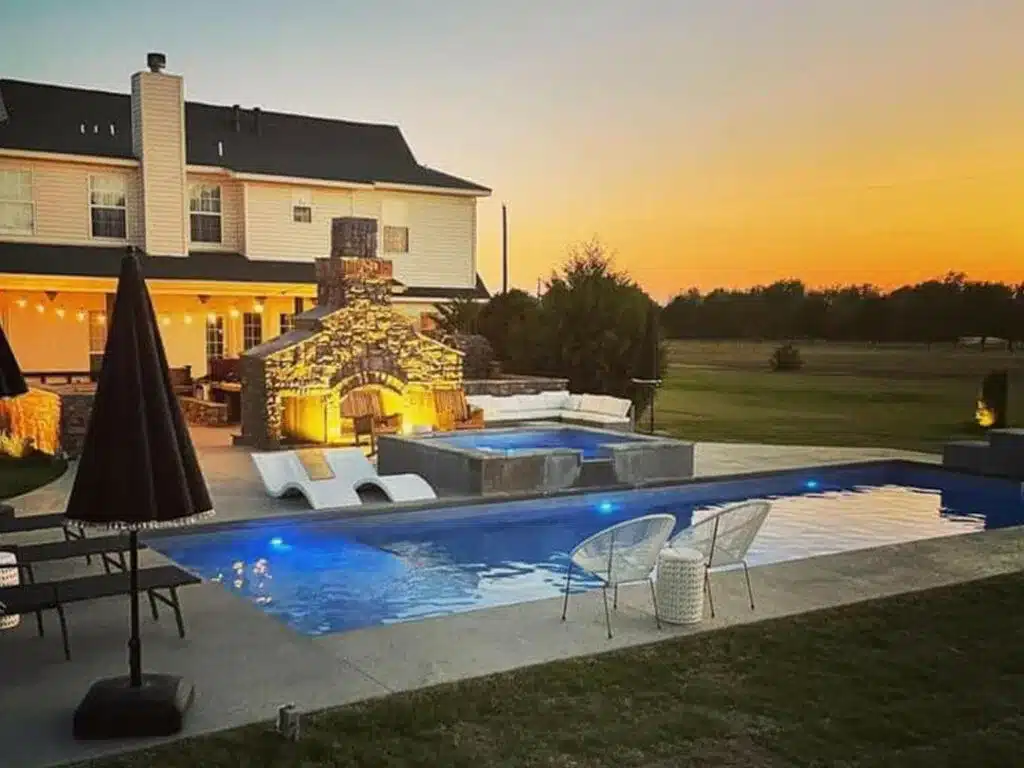 Modern backyard swimming pool with spa, outdoor fireplace, patio seating, and sunset lighting