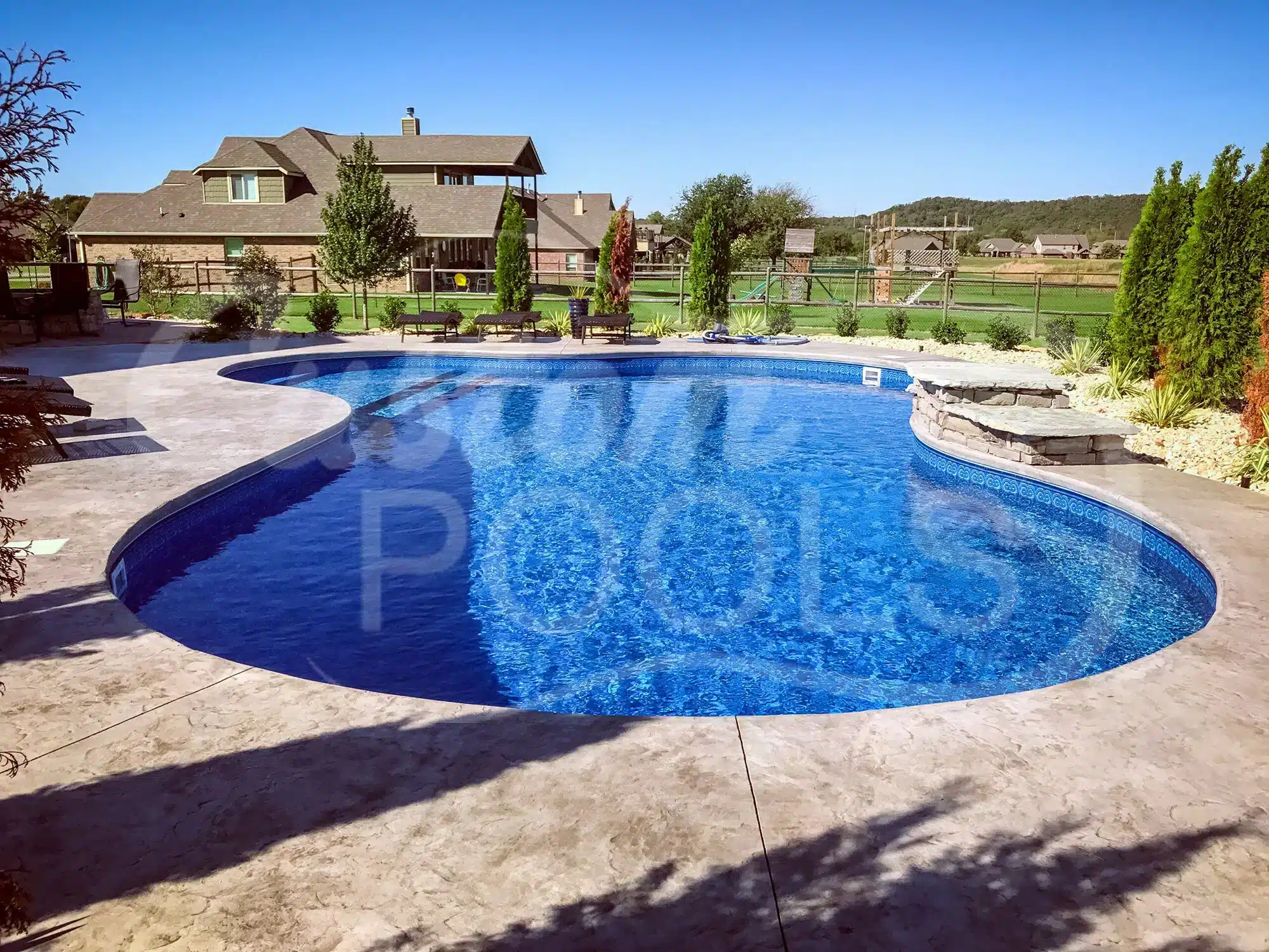 Freeform backyard swimming pool with clear blue water, concrete deck, stone seating area, and landscaped garden