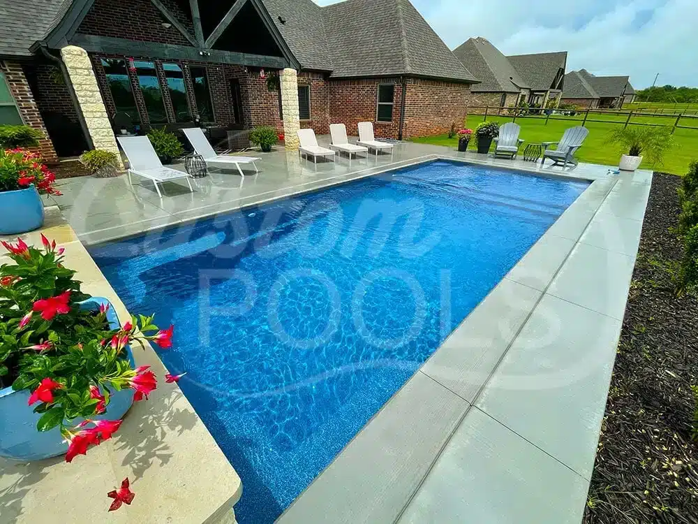 Rectangular backyard swimming pool with blue water, concrete patio, lounge chairs, and brick house exterior