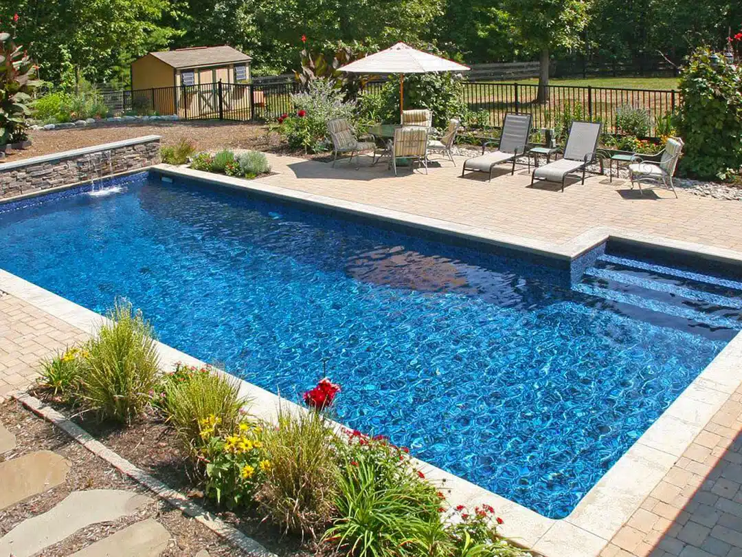 Rectangular backyard swimming pool with blue water, steps, patio seating, umbrella, and landscaped garden